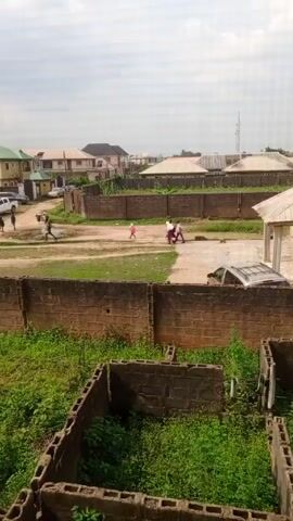 The video depicts a residential area with several houses and a dirt road. The houses are surrounded by brick walls, and there is overgrown greenery in the foreground. Some people are walking along the dirt road, including a group of children. There are also a few parked cars visible. The sky is overcast.