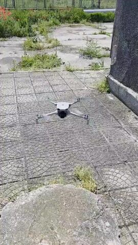 The video shows a small gray drone hovering near a circular stone before landing on top of it. The drone is centered in the frame, and the stone is in the foreground. The background consists of a paved area with cracks and patches of grass, suggesting an outdoor setting. The drone slowly descends and comes to rest on the stone's surface. The video is shot from a static, slightly elevated position, providing a clear view of the drone and its immediate surroundings.