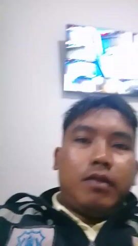 A close-up video shows a man with short, dark hair. He is wearing a dark jacket with a light-colored collar. In the background, a television screen displays an abstract image. The man's facial expression changes throughout the video, from neutral to smiling.