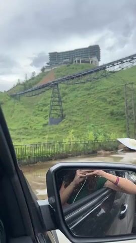 The video shows a drive-by perspective from inside a car. The visible reflection in the side mirror is of the filmer with curly hair, a green top, and a beaded bracelet, holding up hands in a frame-like position. The car moves past a scene of damage and apparent flooding. A large steel truss bridge or conveyor structure spans a green hillside with a building at the top. Below, there is debris, including a flattened white canopy and scattered objects. Houses with balconies are visible on the hillside. The road is covered in water, with additional debris, including pipes and sections of stone wall. The weather appears overcast and rainy.