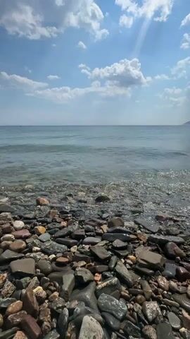 This is a vertical, eye-level, medium shot of a rocky beach on a sunny day. The foreground is filled with an abundance of smooth, varied-colored stones, ranging from dark grey and brown to lighter shades of beige and white, some appearing wet from the receding tide. The middle ground features a gentle wave rolling onto the shore, creating white foam as it washes over the pebbles. The water is a calm, clear blue-grey, reflecting the bright sky. The background shows a vast expanse of the ocean meeting the horizon, with a faint, mountainous coastline visible in the distance on the right. The sky is a vivid blue, dotted with fluffy white clouds, and sunlight beams down from the upper right, casting a bright sheen on the water's surface. The overall mood is peaceful and serene, evoking a sense of a quiet summer day by the water. There is no visible text in the media.