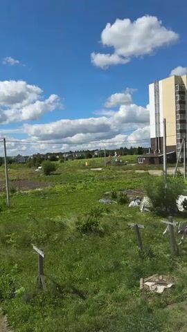 The video shows an outdoor scene on a sunny day with a bright blue sky and scattered white clouds. In the foreground, there is a garden with raised beds, some covered with white tarps and netting. There are also some wooden stakes in the ground. In the middle ground, there is a grassy field with some bushes and trees. A utility pole with wires runs across the scene. In the background, there are houses and more trees. A tall, modern building with a beige and white facade is visible on the right side of the frame. The overall impression is a semi-rural or suburban setting with evidence of both natural landscape and human development. There are no people visible in the video.
