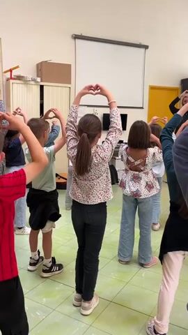 A group of children and an adult are seen from behind, participating in a synchronized movement activity indoors. They are all facing away from the camera, with their arms raised and hands clasped to form a heart shape above their heads. The adult, also facing away, appears to be leading the activity. The setting is a brightly lit room with light green tiled flooring and white walls, typical of a school or community center in La Fouillouse, France. In the background, there is a large white projection screen, a flat-screen television beneath it, and various storage cabinets. A yellow door is visible on the right side of the room. The participants are casually dressed, with children wearing jeans, t-shirts, and floral tops, and the adult in a dark top. The atmosphere suggests an engaging group activity, possibly a dance or exercise session. No visible text is present in the image.