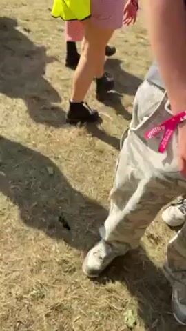 Here's a description of the video:

The video seems to be shot at an outdoor event, possibly a festival, based on the presence of colorful flags, numerous people in the background, and grassy areas. The video begins with a person wearing a gray sleeveless shirt and light-colored pants. 

The camera then moves to show a person in a white tank top holding a red plastic cup, with other people visible in the background. 

Next, the perspective switches, and a person lying on the ground with a blue cap is being assisted by another individual. The person on the ground appears to be in distress, possibly injured or experiencing discomfort.  The scene is surrounded by other people, some sitting at picnic tables in the background.