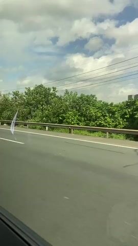 The video shows a white car driving on a highway. The car is in the right lane, and there is greenery and trees on the left side of the road. The sky is partly cloudy. The license plate of the car is partially visible, showing "NL 04992". The video appears to be shot from another vehicle, possibly a passenger in a car traveling in the same direction.