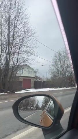 The video captures a view from inside a moving car, looking out the passenger side window and partially into the side mirror. The scene depicts a roadside in a rural or suburban area, identified as Belavino, Russia.

The ground is partially covered with patches of snow, indicating late winter or early spring. The sky is overcast and grey. Bare trees, typical for the season, line the road.

Several houses are visible:
- In the opening shot, a light green two-story house with a red garage door is seen on the left.
- Later, a white house with a distinctive red gambrel roof and a wooden fence is visible.
- Other houses, some red brick, can be seen in the distance.

Various road infrastructure elements are present, including power lines, utility poles, and streetlights. Two prominent road signs are visible:
- A blue pedestrian crossing sign (a square with a white triangle and a black figure walking).
- A "no overtaking" sign (a red circle with two cars, one red and one black, with a diagonal line between them).

Briefly, a helicopter is seen flying in the distance against the cloudy sky. The car's side mirror reflects the road behind and the car's own body, which appears to be a light brown or gold color. The video shows continuous movement along the road.