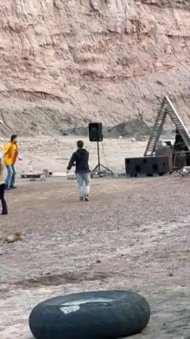 The video shows a group of people dancing in a quarry.  There is a speaker on a stand playing music. Behind the group is a steep quarry wall with visible layers. To the right is an angled ladder leaning against some machinery. In the foreground is a large, worn tire. Most people are standing on the left side of the frame, while two people are dancing near the speaker.  The ground is gravel and dirt. The lighting suggests it is daytime, but somewhat overcast.