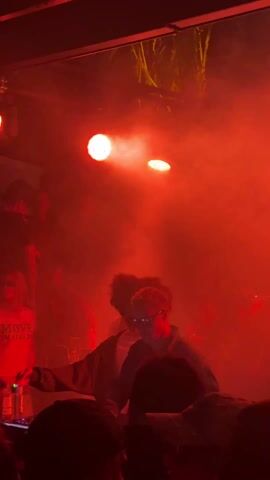 The video captures a DJ performing in a dimly lit club environment, bathed in red light and haze. A DJ with short blonde hair and glasses is seen manipulating the DJ equipment while surrounded by a crowd of people. The crowd appears to be engaged and enjoying the music. Some attendees are facing the DJ, while others are talking among themselves. The scene is further illuminated by round stage lights and faint decorations hang from the ceiling.