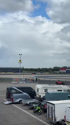 The video shows a parking area with several vehicles, including vans, campers, a trailer, and a motorcycle. In the background, a race track is visible with spectators in the stands. The sky is cloudy.
