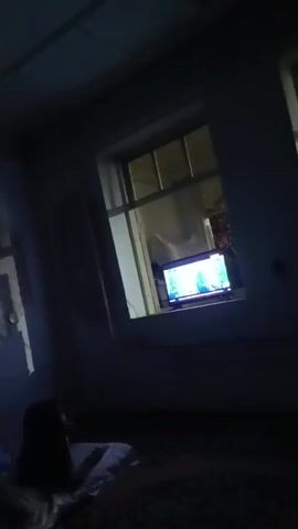 A dark room with a person lying on the floor and a television screen visible through a window. The television displays an image.