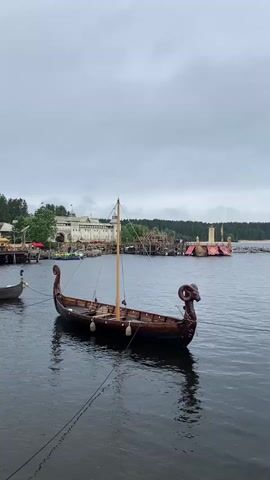 The video shows a view of a body of water with several boats docked. In the foreground, a traditional Viking-style longboat with a ram's head carving on the prow is moored. The boat is made of dark wood and has a single mast. To the left, another smaller boat is visible. In the background, a large, light-colored building resembling a castle or fort is situated on the shore, with several smaller wooden structures and scaffolding around it. Trees line the background. The sky is overcast. There are people visible on the water in the middle ground, near the shore, in what appears to be a blue raft or boat. The overall scene suggests a historical reenactment or themed attraction, possibly a Viking village or a movie set. The water is calm, and the lighting is muted due to the cloudy sky.