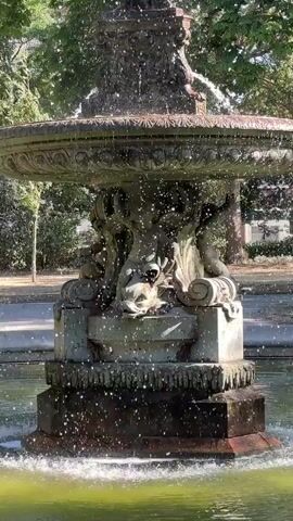 A close-up shot focuses on an ornate, multi-tiered fountain in a park. Water cascades from the top tier, falling onto lower levels and splashing into the pool below. The fountain features intricate sculpted details, including figures and what appears to be a dragon-like creature. Lush green trees and foliage form the background. In the lower left corner of the frame, two people are visible, appearing to walk past the fountain. The sunlight highlights the falling water droplets, creating a shimmering effect.