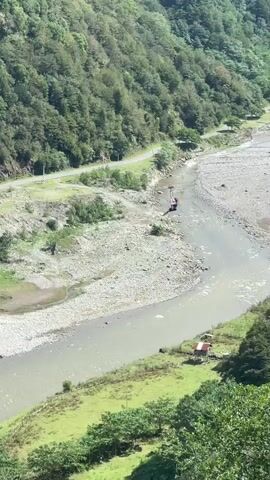 A winding road is visible on a hillside covered with trees. A river flows in the foreground. There are a few small structures along the riverbank and on the hillside. People are visible on the side of the road and near the river. A car is driving on the road.