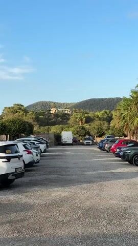 A parking lot filled with vehicles is visible in the foreground. In the background, a large house sits atop a hill surrounded by trees. The sky is clear and blue.
