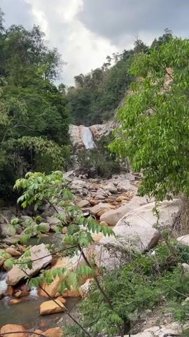 The video shows a rocky riverbed with a waterfall in the distance. The water is flowing over red-brown rocks and around large white boulders. Green trees and vegetation surround the riverbed. The sky is overcast and gray.