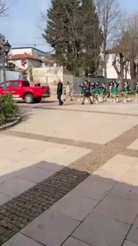 A wide-angle shot captures a parade of children marching along a tiled square on a sunny day. The camera follows the parade from right to left, showing two distinct groups of children. The first group is dressed in green outfits with white accents, and the second group is in red and white. They march in a line and are accompanied by music. People walk on the sidewalk to the left, including two people in black, and a woman in a tan coat. A red pickup truck is parked near a building under construction in the background. The environment includes bare trees and a clear sky.