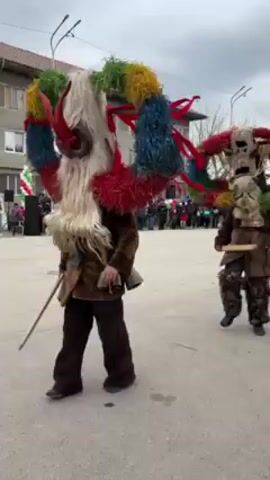 The video shows a group of people dressed in traditional costumes walking down a street. The costumes are elaborate, with colorful and furry masks that extend high above their heads. The people are walking towards a crowd of onlookers. The street is paved and lined with buildings. The sky is overcast.