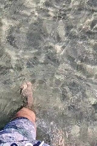 The video begins with a close-up shot of feet wading through shallow, clear water. The camera then pans up to reveal a body of water with a rocky bottom, and further still, a wider view of a beach with boats anchored offshore under a clear blue sky. The water is calm and transparent, revealing the pebbles and sand beneath the surface. Two boats are visible in the distance, one larger and darker, the other smaller and lighter. The scene suggests a peaceful day at the beach or seaside.