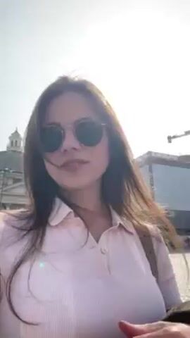 A young woman with long brown hair and wearing a pink polo shirt and round sunglasses films a selfie video in a sunny, outdoor location. Behind her is a tall obelisk with visible inscriptions. Several people are walking around in the background.  The sky is clear and blue.