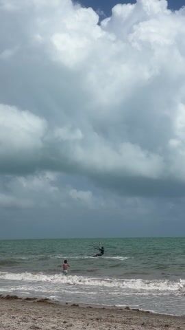 The video shows a beach scene with a body of water, likely the ocean, under a sky that is partly cloudy. The clouds are predominantly white and billowy, with patches of blue sky visible. 

In the water, there are two individuals. One is kite surfing with a teal-colored kite high above, and the other person is standing in the shallows near the shore. The surfer is further out in the water, gliding across the surface. 

The beach is sandy, with some seaweed or debris washed up along the shoreline. The waves are small and gentle. The overall scene is indicative of a recreational beach setting.