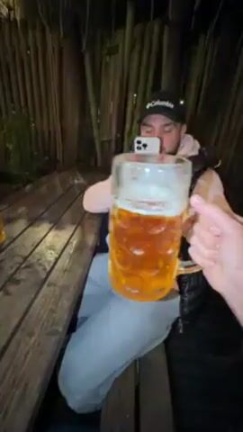 The video shows two people sitting at a wooden picnic table outside at night. One person is holding a large glass beer mug, and the other person uses their phone to record a video of both mugs clinking together. A third beer mug is also visible on the table. The person recording the video is wearing a black cap and a pink sweatshirt under a black vest. A wooden fence is visible in the background.