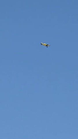 A small yellow airplane is captured in flight against a clear blue sky. The airplane is positioned in the upper right quadrant of the frame and is flying from left to right. Its wings and propeller are visible, and it appears to be a fixed-wing aircraft. The background is entirely the expansive, cloudless sky, suggesting an outdoor daytime setting. No other objects or individuals are visible in the frame.