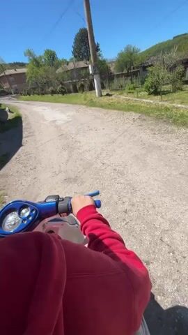 A person wearing a red hoodie and helmet drives a blue scooter down a gravel road. A second person wearing a green vest rides on the back. The road is lined with grass, utility poles, and houses. The sky is clear and blue.