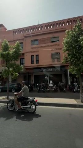 A first-person perspective shot shows a street scene from inside a car. The car is moving, evidenced by the blurred scenery. The street is paved with asphalt, and there are trees and buildings lining the road. In the foreground, a person is riding a motorcycle. The motorcycle is black and has a rider wearing a helmet and a t-shirt. The buildings are a light pink color with ornate window designs. A sign on one of the buildings reads "MONT CRISTAL." Other vehicles, including cars and another motorcycle, are visible on the street. The lighting suggests it is daytime with sunlight casting shadows. The interior of the car is partially visible, including the dashboard and a driver's hand on the gear shift. The overall impression is a view of a bustling street in a city, possibly in North Africa, given the architecture.