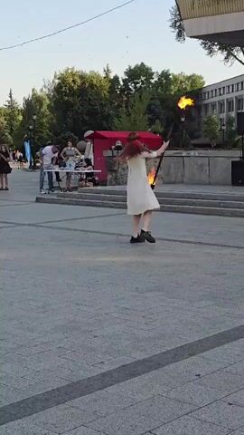 A young woman with long red hair, wearing a light-colored dress and dark shoes, is performing a fire-spinning act in an outdoor public space. She is holding two flaming props, one in each hand, and is in motion, with flames trailing behind her. She appears to be facing away from the camera, moving towards the right side of the frame. In the background, there are several people standing around tables, suggesting a street fair or market. There is also a red tent or canopy, trees, and a modern building with a prominent sign. The ground is paved with large, grey square tiles. The time of day appears to be late afternoon or early evening, given the sunlight and shadows.