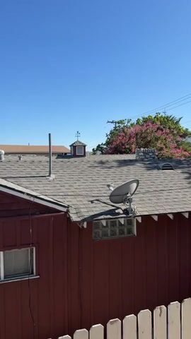 The video shows a close-up view of a rooftop with a blue sky in the background. Several structures are visible on the roof, including a satellite dish, a weather vane on a small cupola, and various vents. A dark red building with a sloped roof is in the foreground, with windows and siding. A white picket fence is visible at the bottom of the frame. The overall scene is an exterior shot of a residential area on a clear day.