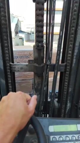 The video is taken from the perspective of someone operating a forklift. The camera is looking forward through the mast of the forklift, which has chains and hydraulic hoses. In the foreground, a hand is visible gripping the steering wheel. In the background, a pallet of boxes wrapped in plastic is visible. Beyond the pallet, there is an outdoor area with a building and a car. The lighting suggests it is daytime and the weather is clear. The forklift appears to be stationary or moving very slowly.