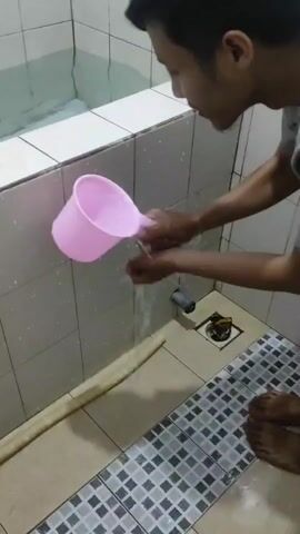 A person is in a bathroom, pouring water from a pink cup onto their face. The person is wearing a gray t-shirt. The bathroom has white tiled walls and a patterned tile floor. There is a bathtub in the background, and a drain on the floor.