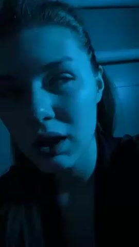 A young woman with her hair pulled back in a ponytail, wearing dark clothing, is seen in a close-up, low-angle shot. She is illuminated by a strong, blue light, giving her skin a blue tint and creating deep shadows. She looks down slightly and moves her lips as if she is talking or singing along to music. The background is dark and out of focus, with only the suggestion of a wall visible.