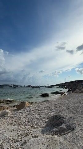 A wide shot of a beach with white pebbles in the foreground. The sea is visible on the left, with small waves lapping at the shore. Several small boats are anchored in the turquoise water. Rocky cliffs form the coastline on the right side of the frame. The sky above is blue with scattered white clouds.