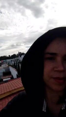 A person wearing a black hooded sweatshirt is seen from the chest up, standing on a red tile roof. The camera is positioned slightly above the person, who is looking directly into the lens with a neutral expression. Part of their face is obscured by the hood. The background includes a residential street, various houses, and a cloudy sky. The neighborhood appears suburban.