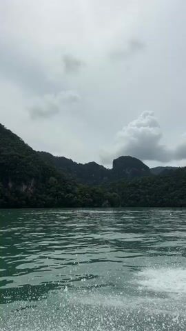 This is a first-person perspective video taken from a moving boat. The boat is creating a wake in the turquoise water, which is visible in the foreground. In the mid-ground and background, there are lush green, tree-covered mountains. The sky is cloudy and overcast. The boat appears to be traveling along the coast of an island or a mountainous region. The overall mood of the video is serene and natural.
