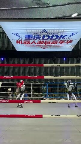 The video shows two humanoid robots in a boxing ring. One robot is wearing red boxing gloves and is on the left side of the ring. The other robot is on the right side of the ring and is wearing white boxing gloves. The robots are facing each other and appear to be in a fighting stance. A large screen above the ring displays Chinese characters and a logo. The boxing ring has red and white ropes and is set up in a large venue with spectators visible in the background. The lighting in the venue is dim, with spotlights illuminating the ring.