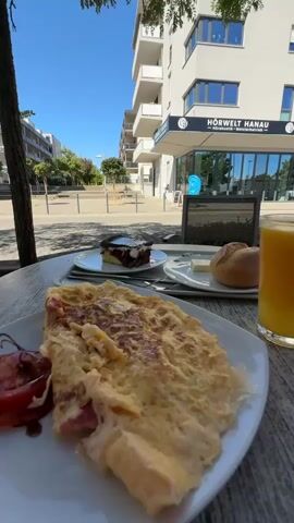 The video begins with a close-up of an omelet on a plate, accompanied by a slice of tomato. In the background, a slice of cake with cherry and cream is visible, along with a bun and butter, and a glass of orange juice. The scene is set at an outdoor cafe with modern buildings in the background. The camera then pans to the left, showing more of the street and buildings. Next, the camera focuses on a young woman with dark curly hair, wearing a dark-colored top and white earbuds, as she looks at the camera. The background shows a building with a sign that reads "Wiener Feinbäcker." The video then quickly pans to the right, showing blurred outdoor seating and foliage. The final shot is blurry and depicts outdoor seating and greenery.