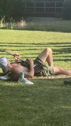 A person is lying on their back in a grassy field, playing a guitar. They are wearing a white baseball cap, green camouflage shorts, and no shirt. A blue and white can is next to them, along with a pair of white sneakers. There is another person lying down further to the right, partially obscured by a shadow. In the background, there are trees, a body of water, and some modern buildings. The scene is lit by sunlight, casting shadows on the grass.