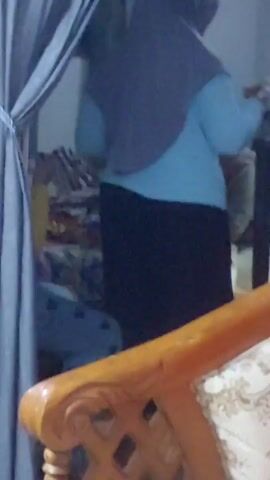 A person wearing a blue long-sleeved shirt and a dark hijab is standing with their back to the camera. They are wearing a black skirt. The person appears to be near a window with blue curtains. Another person is partially visible in the background. The image quality is low and blurry.