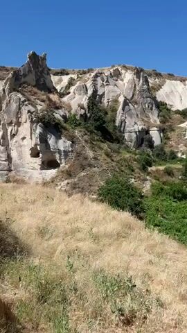 The video displays a landscape with unique rock formations, characterized by their pale, layered appearance and natural erosion. These formations have a somewhat conical or columnar shape in places, with several appearing to be carved out or naturally hollowed. Lush green vegetation, including trees and shrubs, grows at the base of these rock formations and in the foreground. The ground is covered with dry, yellowish grass. The sky is a clear, bright blue, indicating a sunny day. The camera is panning slightly to the right, revealing more of the rocky landscape and the surrounding vegetation.