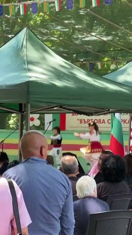 A group of people wearing traditional Bulgarian clothing are performing a folk dance on a stage under a green canopy. The stage is decorated with Bulgarian and European Union flags, as well as a banner that reads "Българска Сърце" (Bulgarian Heart). There are two young boys and two young girls dancing, and they are all wearing brightly colored costumes. The boys are wearing white shirts, black vests, and white trousers, while the girls are wearing white blouses, colorful skirts, and embroidered aprons. An audience is seated in front of the stage, watching the performance. The scene is outdoors, and there are trees visible in the background.