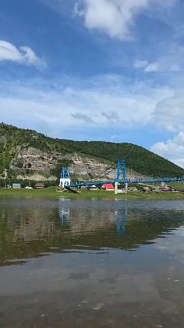 The video shows a wide river with a blue bridge spanning across it in the distance. The bridge has two towers and is supported by cables. On the other side of the river, there is a village with several houses. Behind the village, a large, green, mountainous landscape rises up. The sky is blue with some white clouds. The camera angle is low, looking across the water towards the bridge and the landscape. The water's surface is calm and reflects the sky and the landscape. There are no people visible in the video.