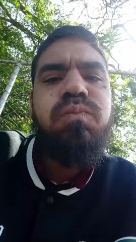 The video shows a close-up shot of a man with a beard exhaling vapor. He is wearing a black jacket with white trim around the collar. Behind him is a chain-link fence and trees. The sun is shining through the trees, creating dappled lighting. The camera angle is low, looking up at the man's face. He looks directly into the camera.