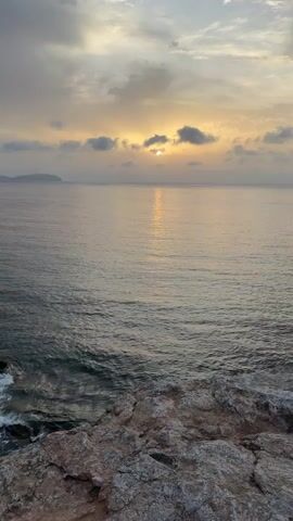 This is a vertical, eye-level, long shot video of a sun setting over a calm ocean. The sun is partially obscured by clouds, casting a golden reflection on the water. The foreground is dominated by a rocky cliff face. The sky is a mix of gray and yellow clouds. There are no people or animals visible.