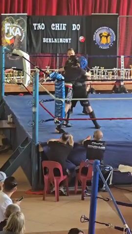Here's a description of the video:

The video shows two kickboxers in a ring, engaged in a match. Both are wearing protective gear including headgear, shin guards, and gloves. They are both wearing dark shorts and tops. A referee in black stands nearby, observing the fight. 

The ring is surrounded by ropes and turnbuckles. Spectators are seated around the ring, and some are watching the fight intently. One man is sitting with "Coach Bensihmed" printed on his shirt.

Behind the ring are banners displaying the name of the event and affiliations, with trophies displayed on a table. The background includes curtains in red.
