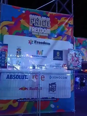 The video shows an outdoor event. The event features a "Maspalomas Pride Freedom Gran Canaria" banner with sponsor logos including Absolut, Red Bull, and RTVE. People are walking around in a crowded area with shops, bars, and rainbow flags strung overhead. There is a DJ or performer visible behind a stage area. The time of day appears to be evening or night.