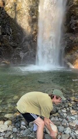 A young man with curly brown hair wearing a light green t-shirt, black athletic shorts with white stripes, and a green baseball cap is standing on a rocky shore next to a waterfall. He is holding a medium-sized, light-colored rock. He tosses the rock into the water, creating a splash. He then turns to face the camera with a surprised or excited expression, his mouth open. The waterfall is powerful, with water cascading down a rocky cliff face. There is a rainbow visible in the mist of the waterfall. The water in the pool at the base of the waterfall is clear and green. The sun is shining, casting shadows from trees on the rocky embankment.