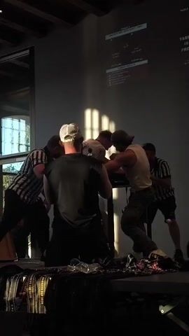 Two men are arm wrestling at a table in front of a projection screen. They are both facing the table with their arms locked. Two referees in striped shirts are standing on either side of the arm wrestlers. There are spectators in the background. The room is dimly lit with sunlight streaming through windows.