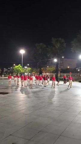At night, under streetlights illuminating a tiled public square, a group of about twenty people wearing matching red shirts and light-colored shorts are rehearsing a choreographed dance. They stand in rows and columns, and move together in a coordinated fashion. In the background are dark trees, buildings and a motorcycle parked to the side.